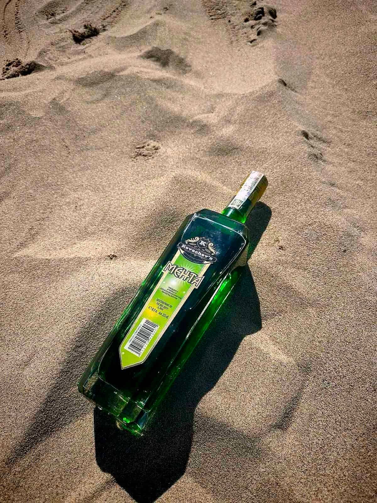 Green bottle of Russian vodka partially buried in sandy beach sand.