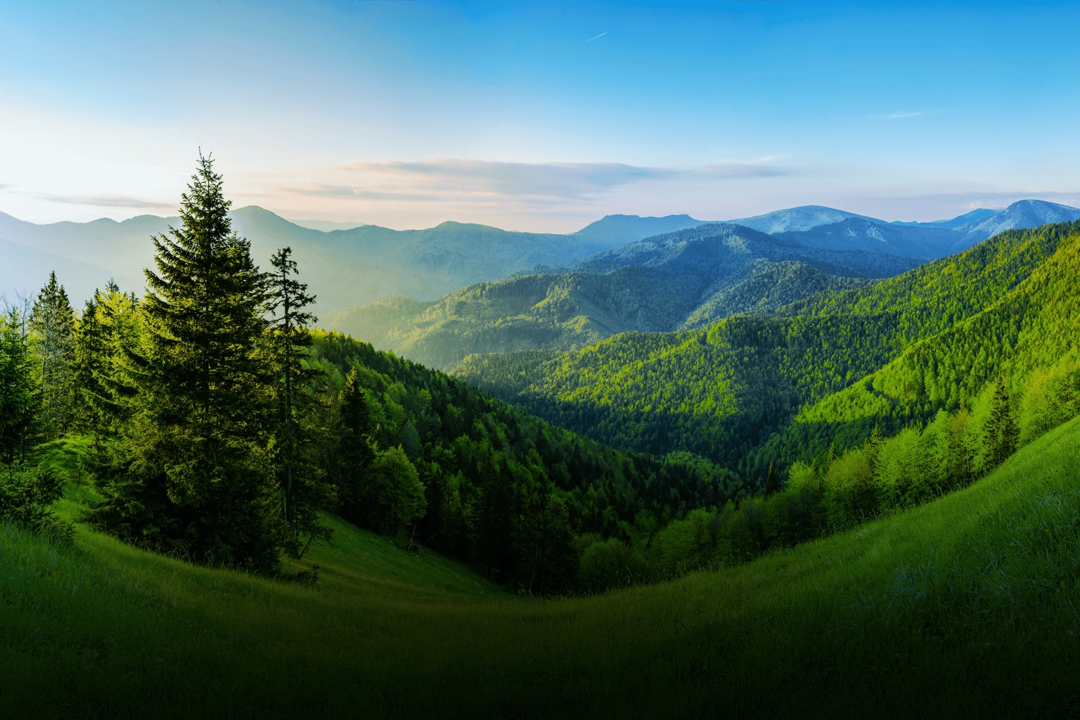 Lush green mountain landscape with dense forests and rolling hills under a clear blue sky.